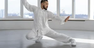 גבר עם זקן לבוש בבגדי טאי צ'י לבנים מסורתיים מבצע תנועת אמנות לחימה בסטודיו מואר. הוא נמצא בתנוחה נמוכה עם רגל אחת מושטת הצידה, יד אחת מורמת ויד שנייה מושטת קדימה, על רקע חלונות גדולים המשקיפים לנוף עירוני.